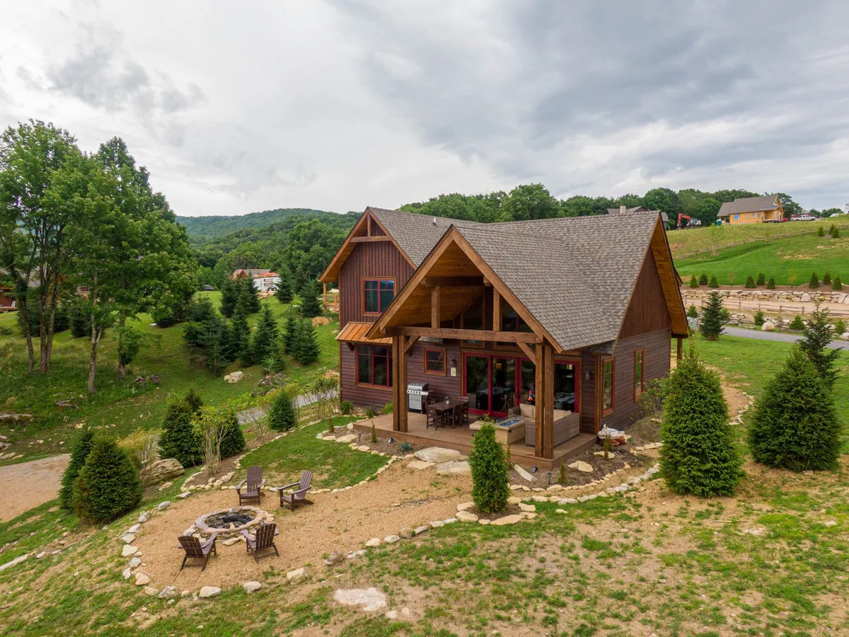 This exterior shot features the house with a spacious porch, a welcoming fire pit area, and abundant greenery. It captures the serene, private atmosphere and offers visual cues for outdoor activities like meals on the porch and nature walks.