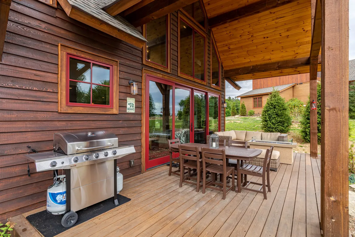 An exterior deck scene featuring a modern grill, an outdoor dining set, and large windows revealing the lush green landscape beyond. The setup invites barbecuing, al fresco dining, and leisurely gatherings under the open sky.