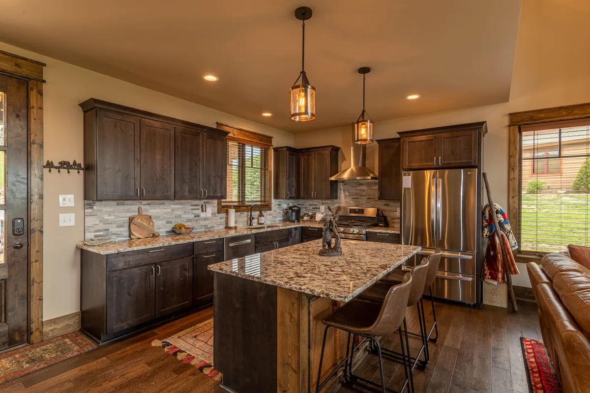 A warm, welcoming kitchen with a prominent granite island, wood cabinetry, hanging light fixtures, and sleek stainless steel appliances. The well-lit space encourages both cooking and conversation, and the island is ideal for meal prep or gathering with guests.