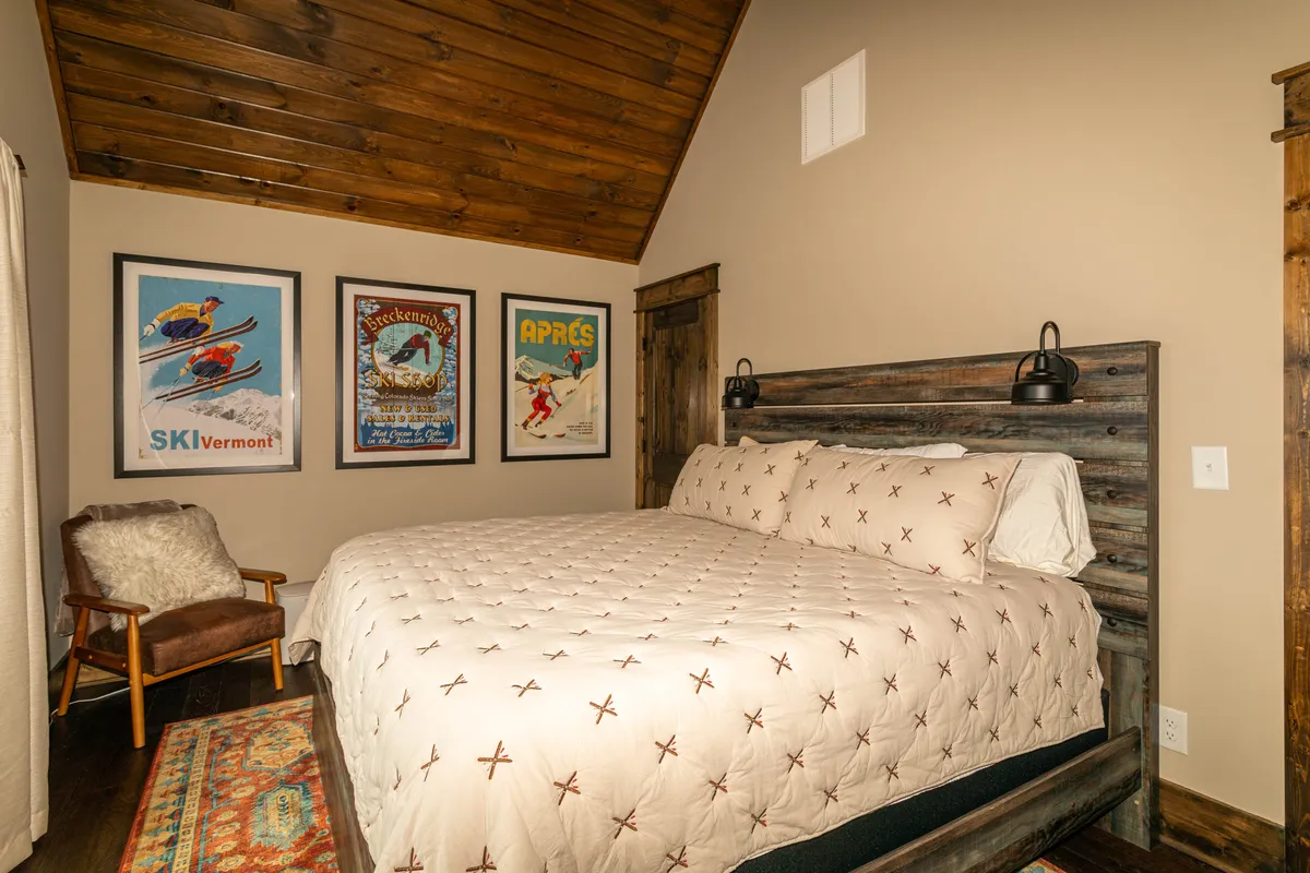 A cozy bedroom featuring a bed with a quilted cover, vintage ski posters on the wall, wooden ceiling, and an inviting armchair. Natural light filters through a window, enhancing the warm, comfortable atmosphere.