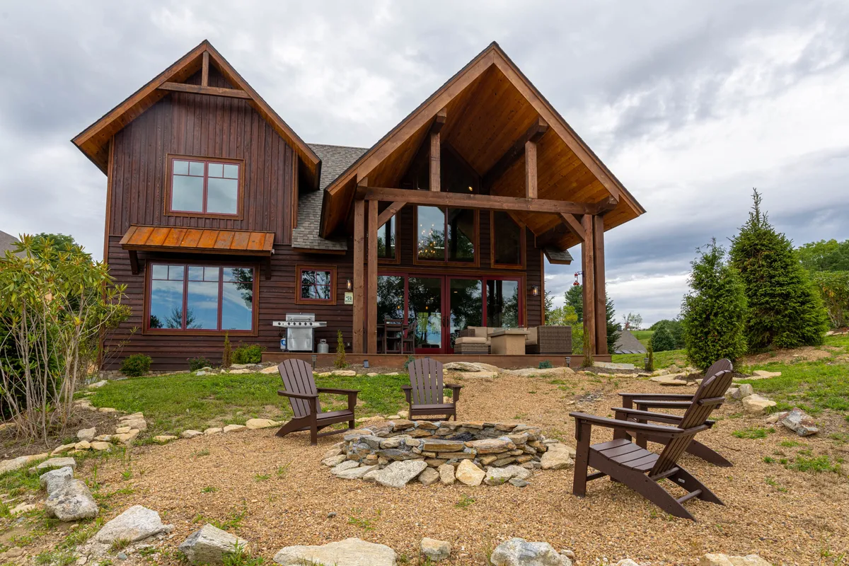 This exterior image showcases a modern cabin with distinctive architecture, a cozy fire pit area, and inviting outdoor seating, all set against a mountain backdrop. The arrangement hints at opportunities for socializing, relaxing, and enjoying natural surroundings.
