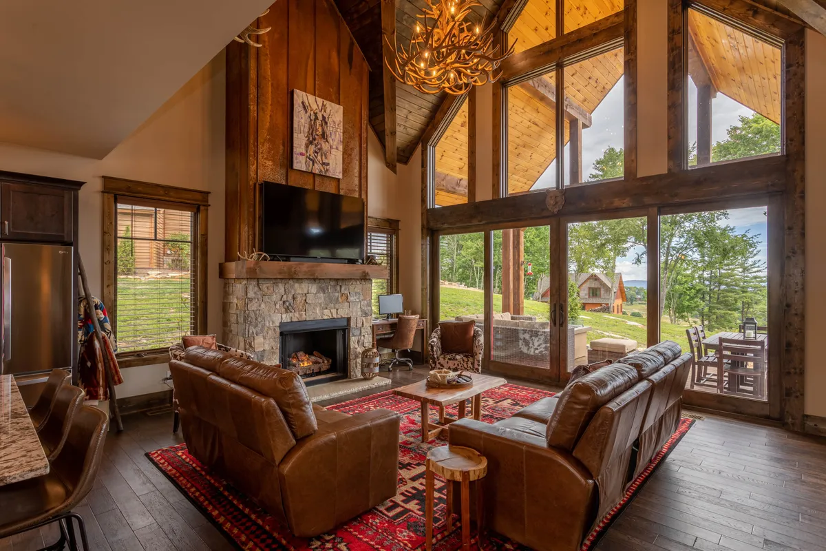A spacious living room featuring a grand stone fireplace flanked by large windows that offer panoramic mountain views. Plush leather sofas are arranged to create a cozy, inviting gathering spot under a rustic chandelier. The room is filled with natural light, and the ambiance suggests comfort for both relaxation and entertaining.