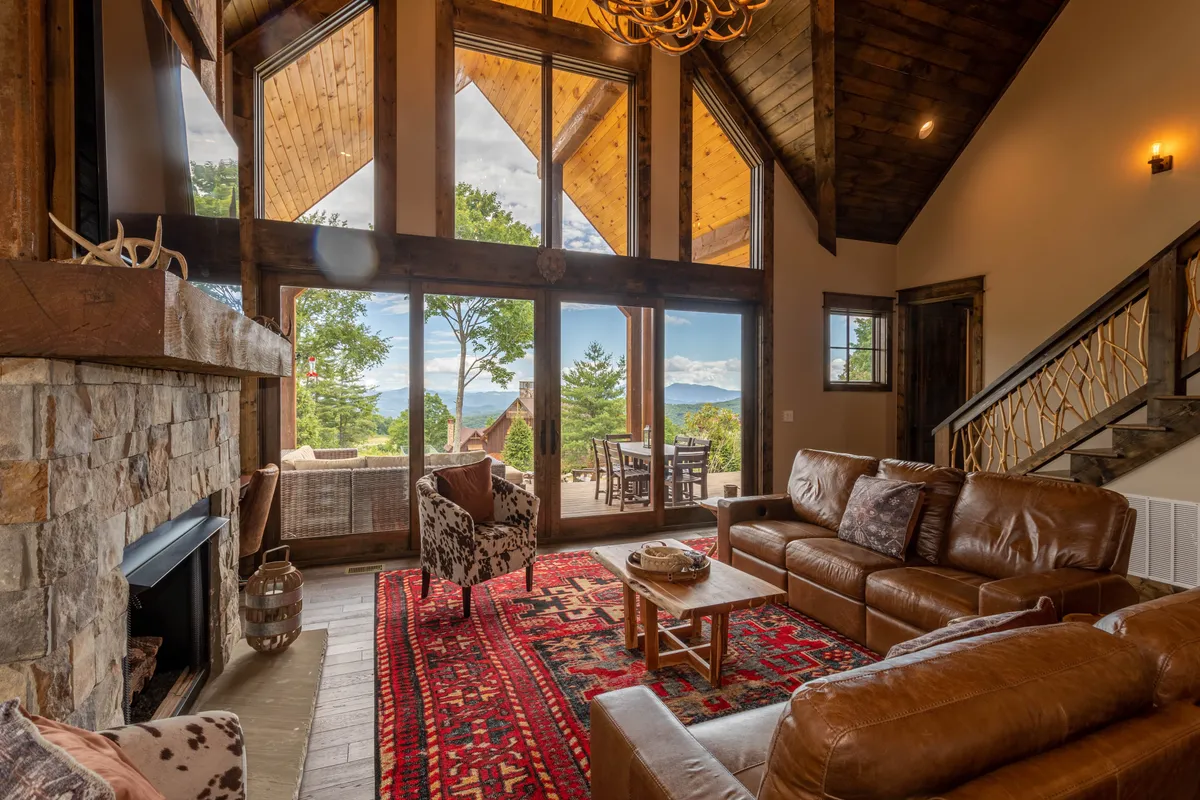 A warm and welcoming living area with a stone fireplace, rustic furniture, a colorful area rug, and large windows that invite in sunset light and mountain scenery. The room feels lived-in and ideal for both quiet relaxation and family gatherings.