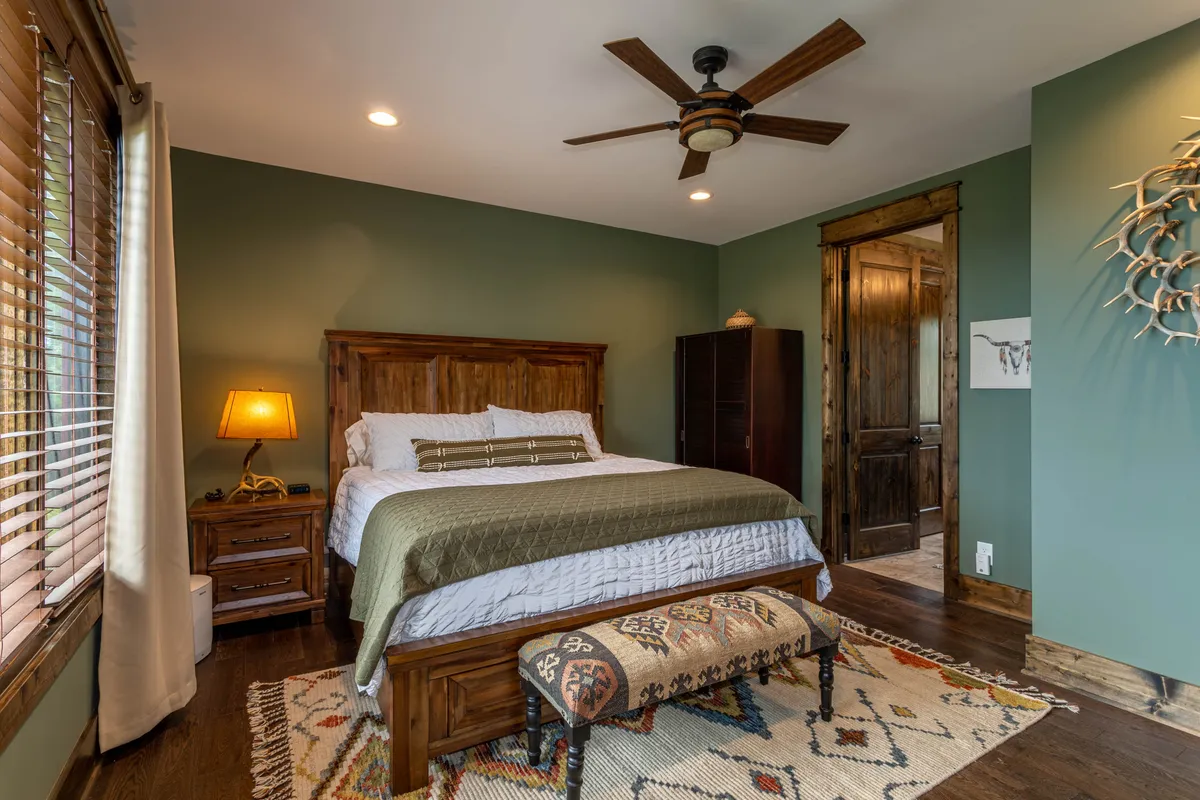 A warmly lit bedroom with a plush bed, nightstand and lamp, soft rug, ceiling fan, and a wooden door. Natural light filters in, creating a homey and restful atmosphere.