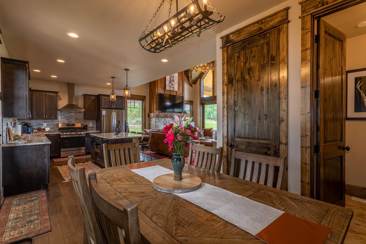 An inviting dining area centered around a table with a floral centerpiece, adjacent to a granite-topped kitchen island and surrounded by warm wood cabinetry. The chandelier lighting enhances the homely, communal atmosphere ideal for memorable meals.