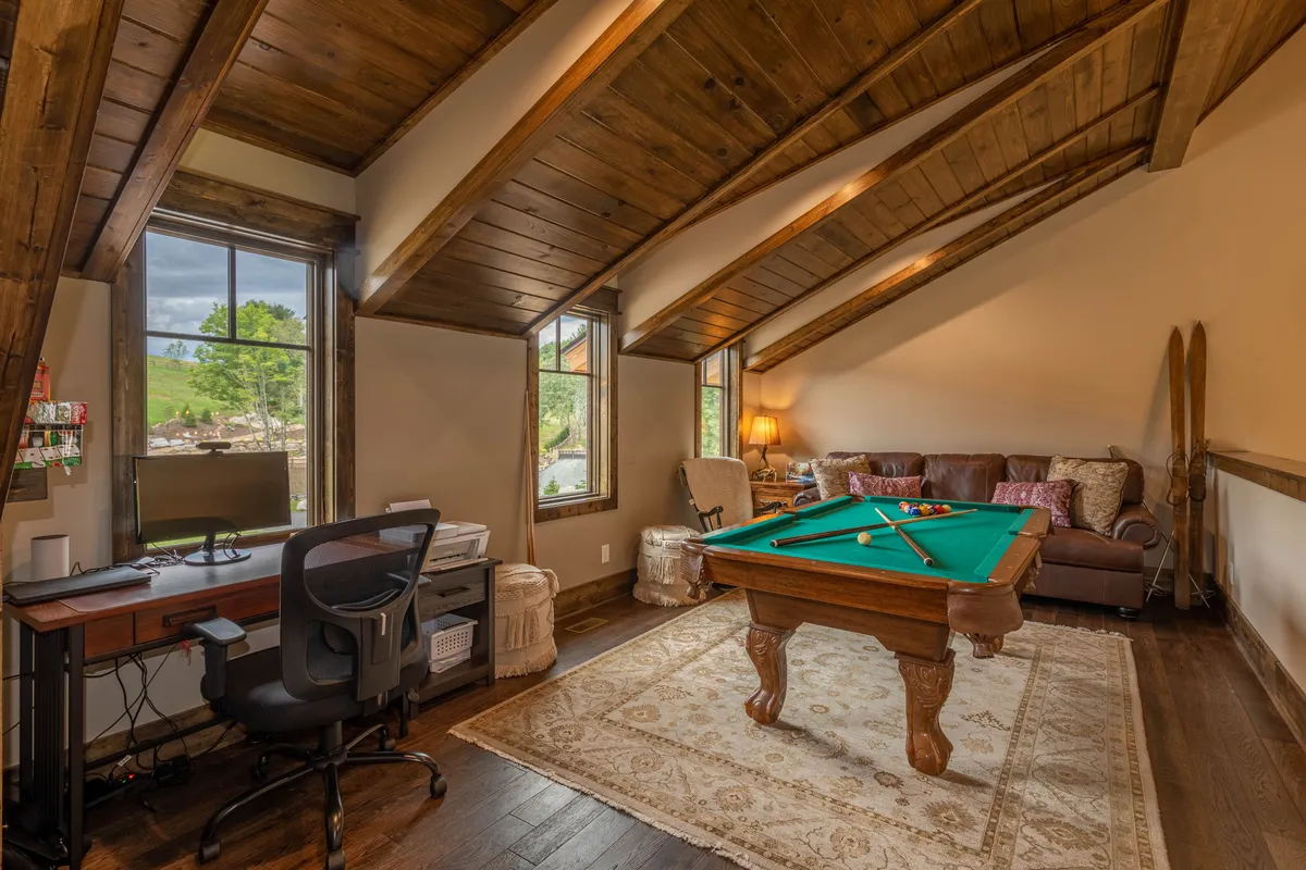 A relaxed loft area featuring a pool table, comfortable couch, wooden beams overhead, and a small desk with a computer. The airy space is designed for play and extra guests.