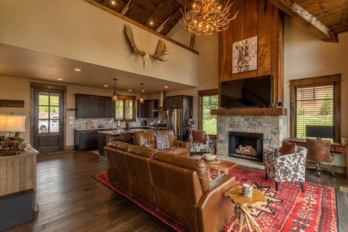 A cozy living space with a large leather sofa facing a rustic stone fireplace, illuminated by a chandelier and accented with wooden beams and artwork. The room has a comfortable, productive atmosphere suitable for work or relaxation.