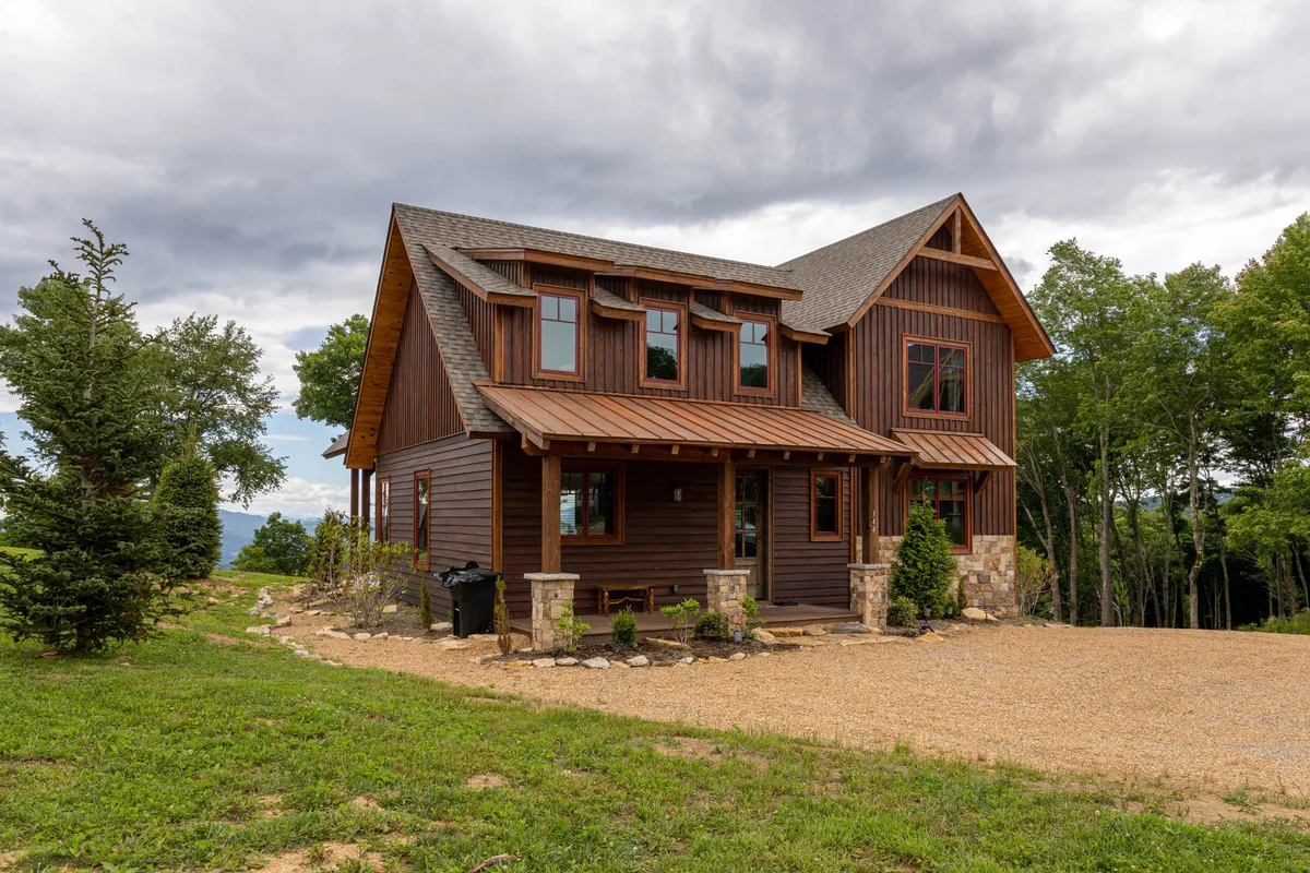 An inviting exterior view of a home’s front porch, showcasing large windows and natural stone accents. The porch is designed for comfortable outdoor seating, surrounded by serene natural beauty, and positioned to capture sunset views.