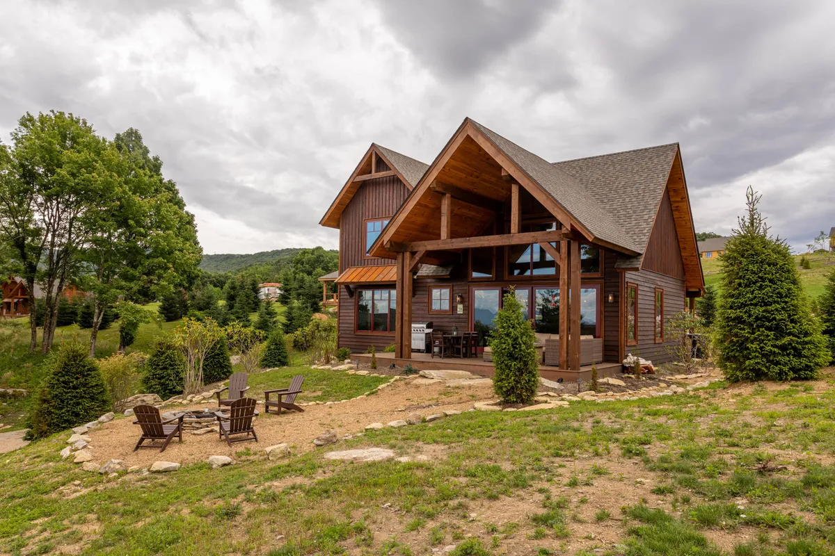The image showcases a newly built mountain cabin surrounded by lush greenery, with an inviting outdoor seating area and a cozy fire pit. The porch offers sweeping long-range views of the Blue Ridge Mountains, perfect for stargazing or sipping coffee at sunrise. The tranquil setting suggests space for gatherings and relaxation in nature.