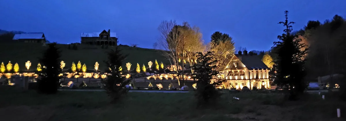 An illuminated exterior view of the house at dusk, with decorated trees and strategic landscape lighting creating a warm, inviting atmosphere. The tranquil scene hints at magical evening strolls or relaxing outside after a day of exploring with your dog. The house's entrance and the surrounding natural area are beautifully highlighted, tying the property to its mountain environment.