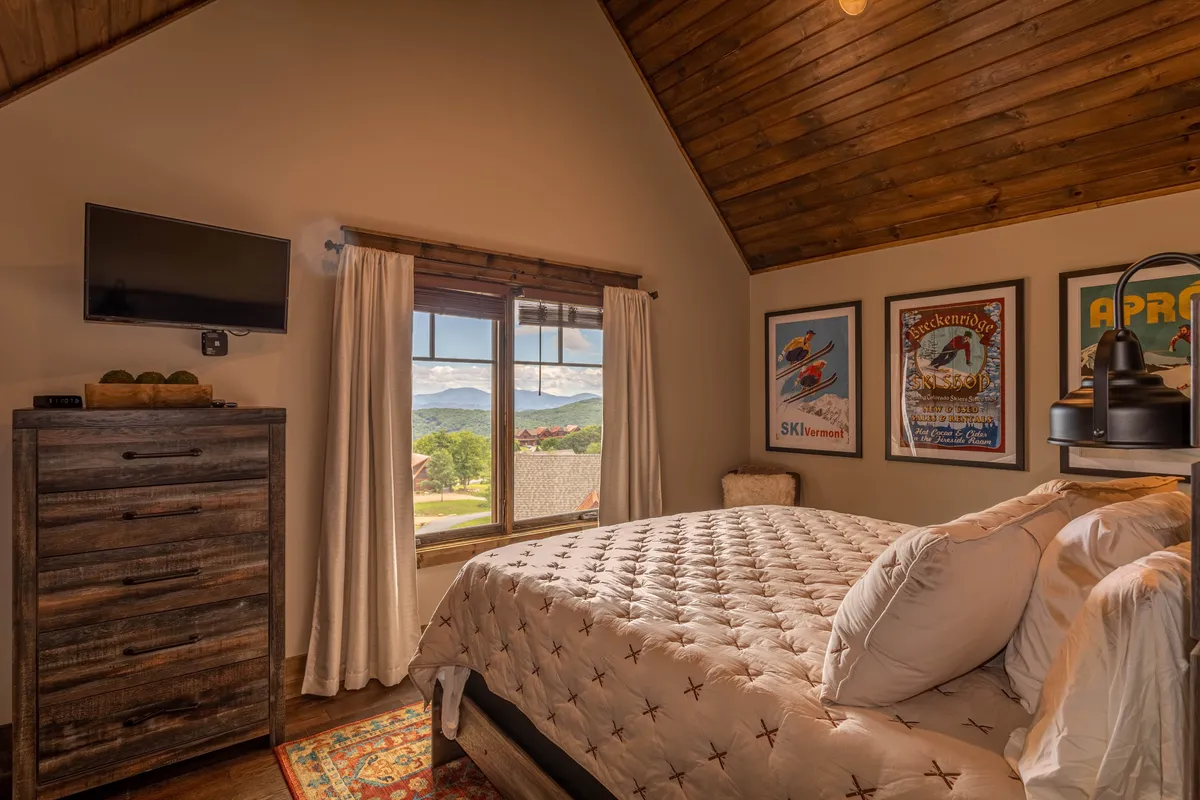 A tranquil bedroom featuring a bed with a quilted cover, large window showcasing mountain views, wall art, and a dresser. The space is serene and bathed in natural light.